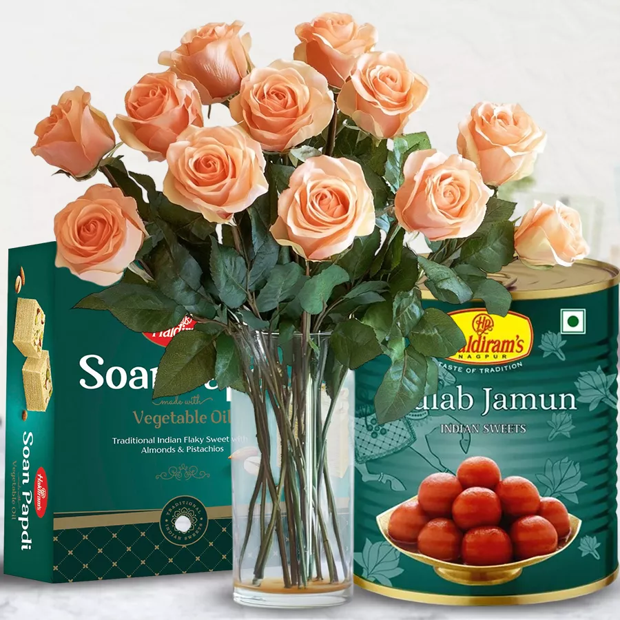 A beautiful arrangement of peach roses in a vase accompanied by Haldiram sweets, including Gulab Jamun and Sohan-Papdi.