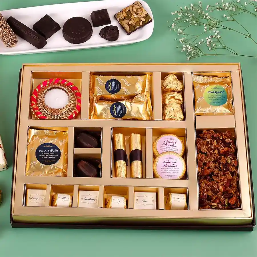 Assorted Delights gift box with Diya, featuring various sweets and chocolates including almond fingers, chocolate rocks, seeds square, brittle, soft-filled chocolates, granola logs, florentines, assorted chocolates, granola, and a traditional Diya.