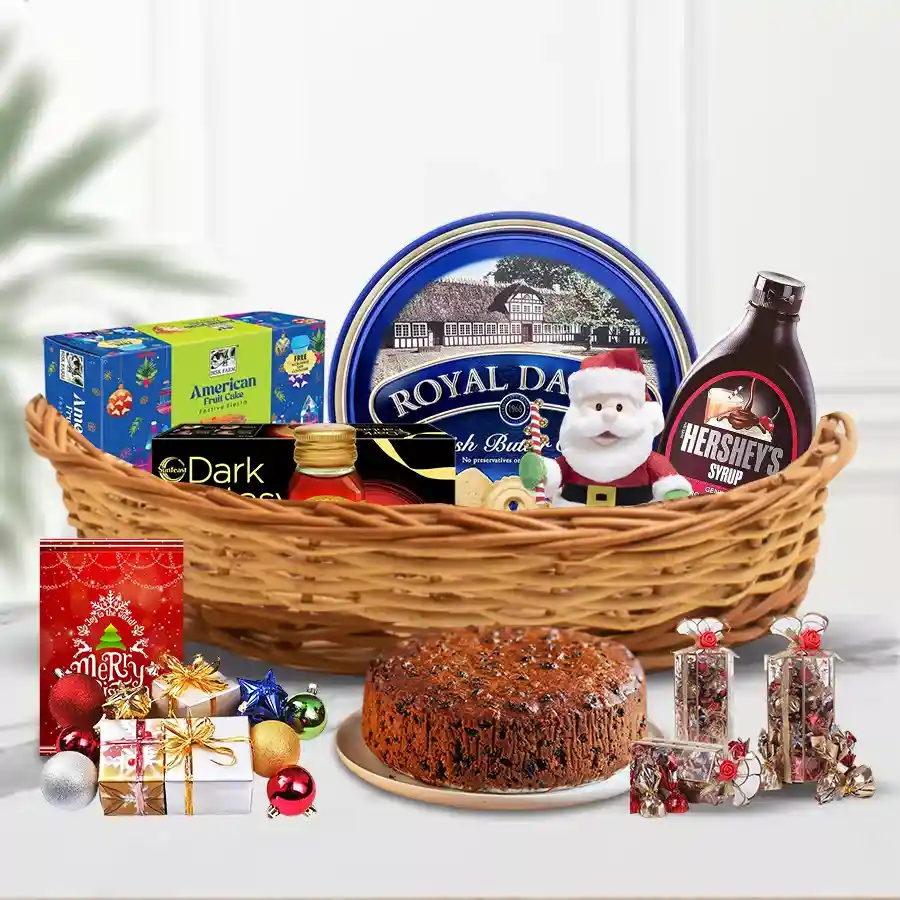 A beautifully arranged Christmas hamper filled with a fruit cake, biscuits, honey, Danish cookies, chocolate syrup, handmade chocolates, a soft toy, a Christmas card, and sparkling decorations in a charming cane basket.