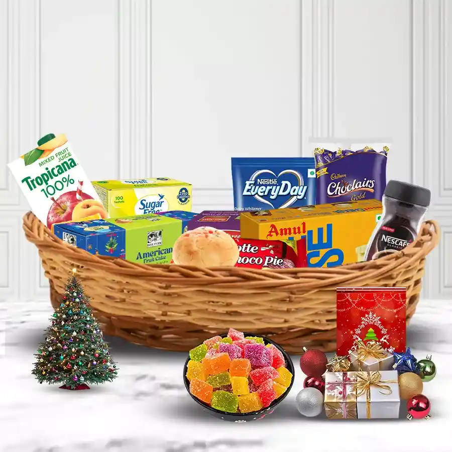 A beautifully arranged cane basket filled with festive treats including American Fruit Cake, Bakerstreet Cookies, Amul Cheese, Nescafe, Tropicana Juice, Lote Choco Pie, Eclairs Chocolates, sweet buns, sugar-free goodies, a small Christmas tree, decorations, a card, and milk powder.