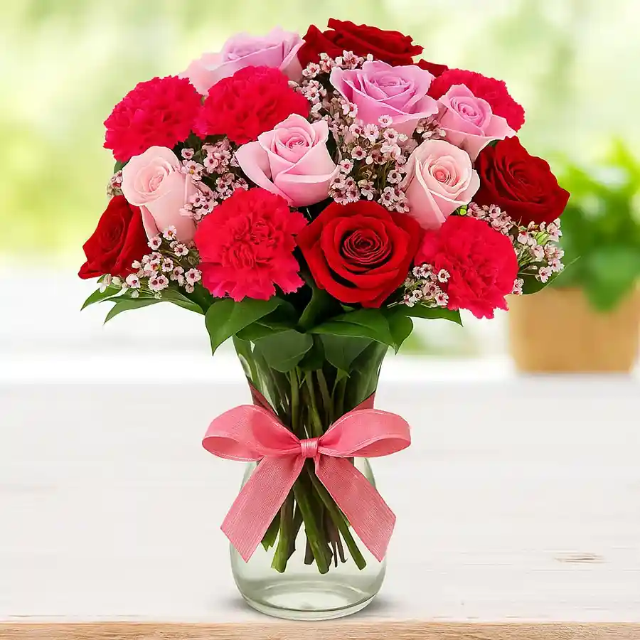 A mixed flowers bouquet featuring red and pink roses and carnations in a glass vase, beautifully arranged and tied with a ribbon.