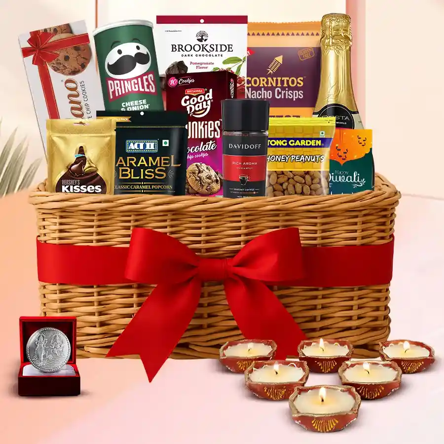A beautifully arranged Diwali gifts basket filled with sparkling fruit juice, caramel popcorn, coffee, dark chocolates, Hershey’s Kisses, assorted cookies, designer mud diyas, a silver-plated Ganesh Laxmi coin, and a decorative cane basket, accompanied by a free Diwali card.
