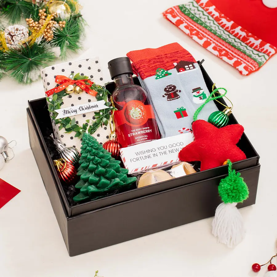 A festive holiday gift hamper filled with a tree-shaped candle, strawberry shower gel, Christmas socks, fortune cookies, a star ornament, and a Christmas activity kit in a colorful confetti box.