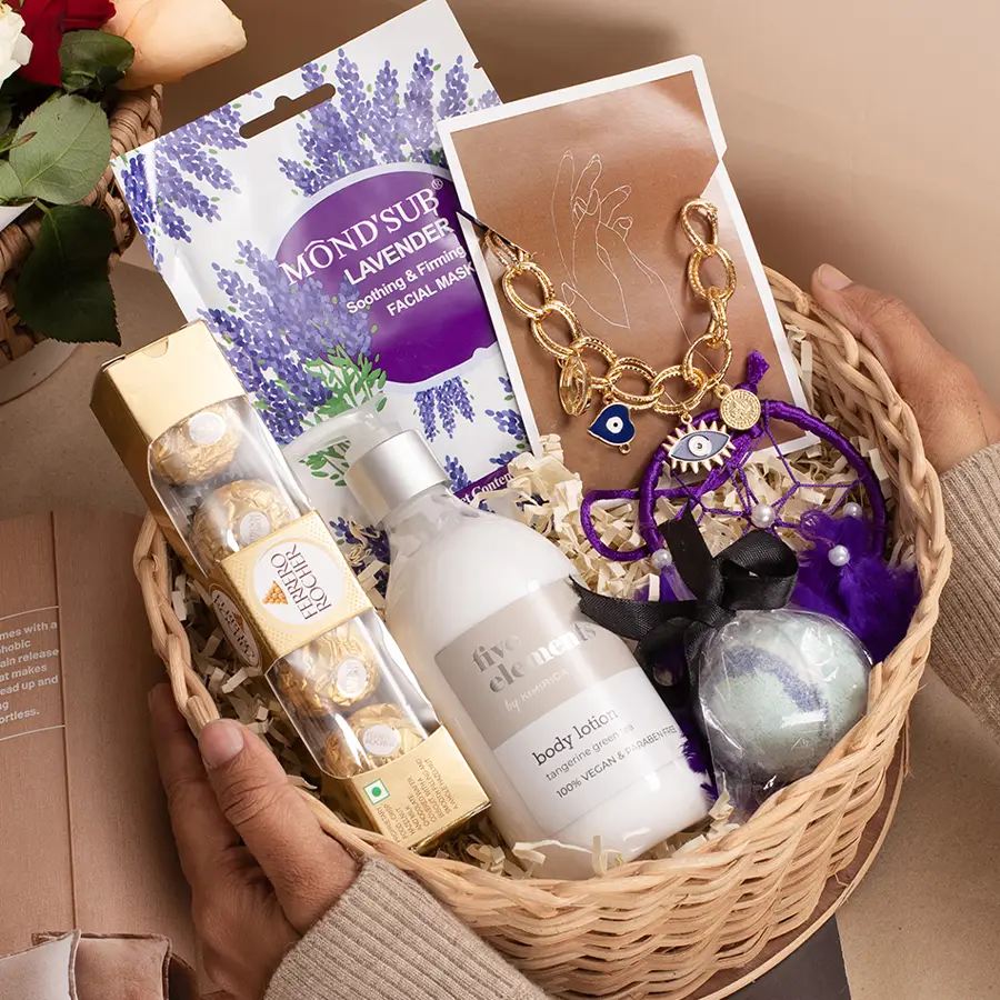 A beautifully arranged gift box featuring a variety of wellness items including an Evil Eye Charm Bracelet, Five Elements Body Lotion, a Purple Dream Catcher, a bath bomb, a Lavender Face Mask, Ferrero Rocher chocolates, and a reusable cane basket.